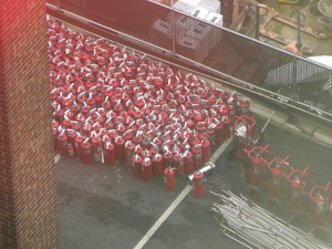 Photo from an upper floor of a hotel, showing about 100 large
red fire extinguishers clustered together in a parking lot. There's a brick
wall along the left of the photo, a construction site in the back (separated
from the lot by a chain link fence with vertical plastic strips), and a big
pile of shiny metal pipes that might belong to a sprinkler system.