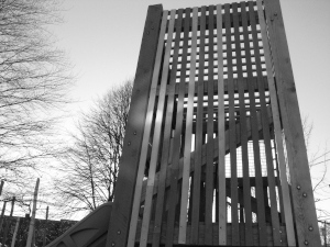 Photo of some tall playground structure, faced with vertical wooden slats.
There are trees against sky in the background.